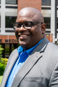 African American man wearing glasses, a blue button-down shirt and gray sports jacket is smiling at the camera.