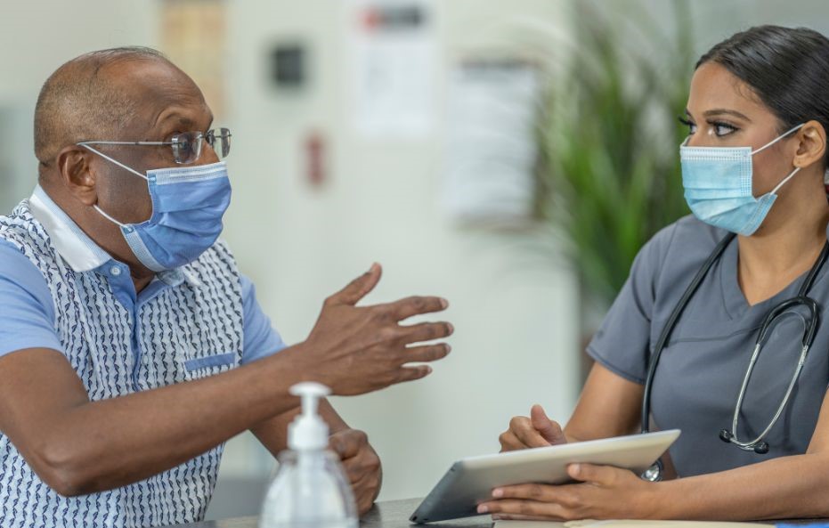 Middle-aged black male patient wearing a mask talks with a young female healthcare worker, also masked