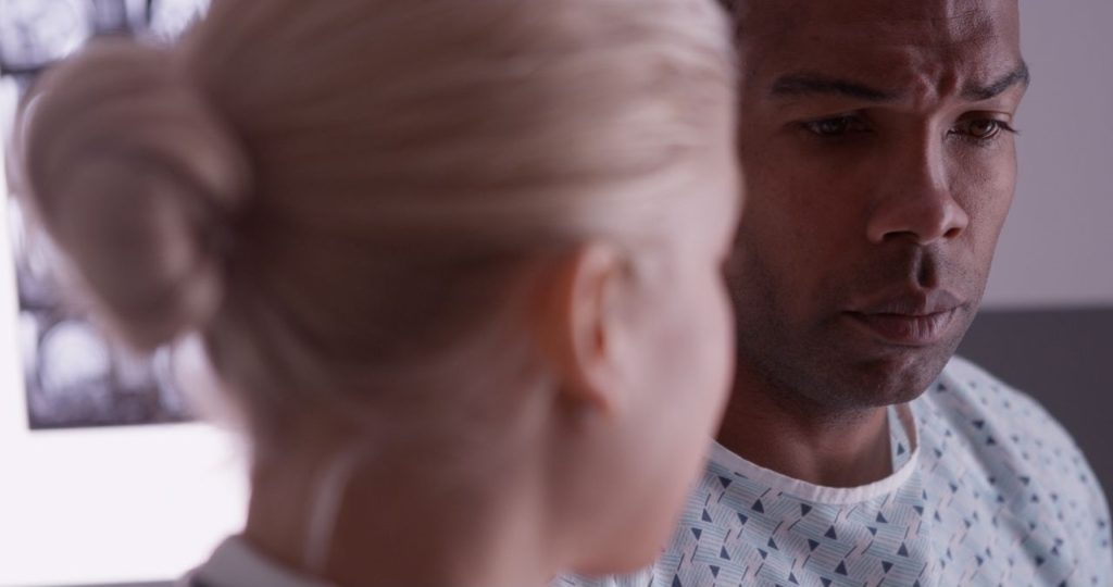 A Black male patient in a hospital gown faces the camera looking unhappy. A white female healthcare provider faces him.