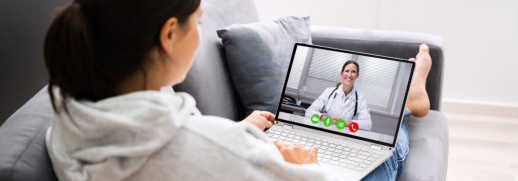 Woman on a couch is interacting with her doctor on a laptop computer.