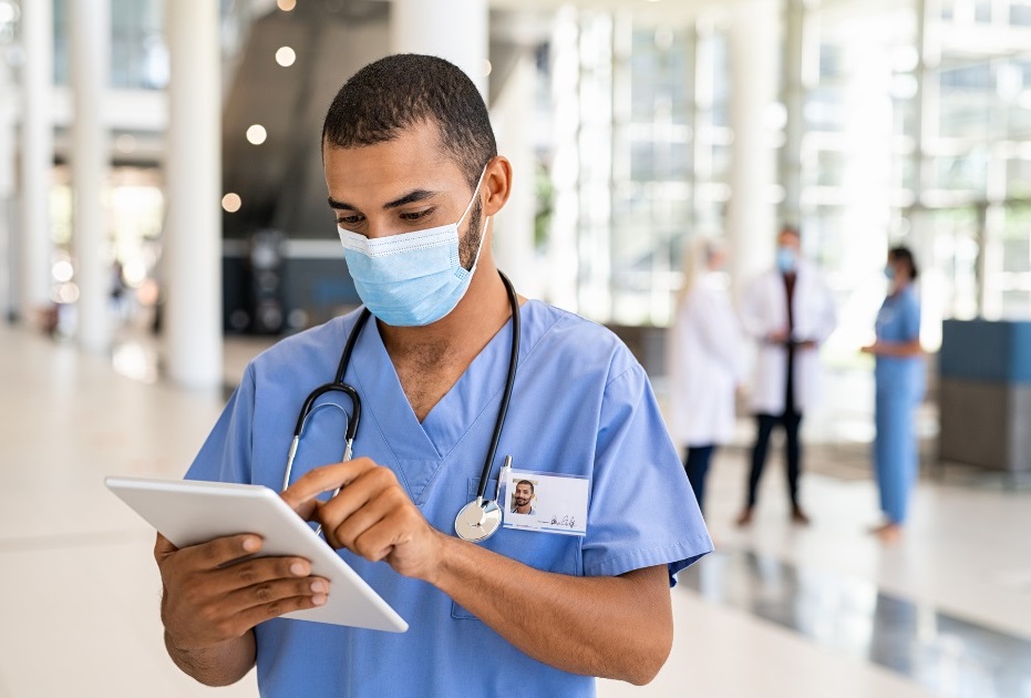 young male healthcare worker standing in a lobby wearing a surgical mask looking at a tablet.